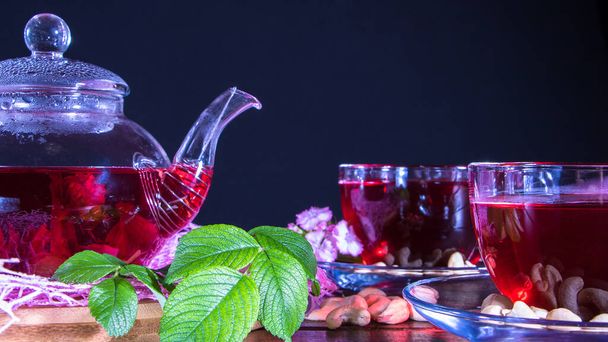 Eine Tasse roten Tee und eine Teekanne mit weißen Hibiskusblüten und grünen Blättern medizinischen Tees auf einem hölzernen Stand.Zen-Teezeremonie. Foto von roten Kräutern indischen Heiltees. Elegante Tassen mit einem entspannenden Getränk - Foto, Bild