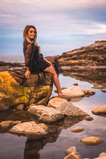 Summer lifestyle with a young brunette Caucasian woman in a long black transparent dress on some rocks near the sea on a summer afternoon. Sitting on top of a rock in a natural pool - Fotoğraf, Görsel