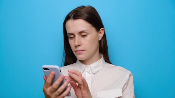 Angry brunette young girl talking with boyfriend, using mobile cell phone, dressed in white shirt, isolated over blue studio background with copy space. People sincere emotions lifestyle concept - Footage, Video