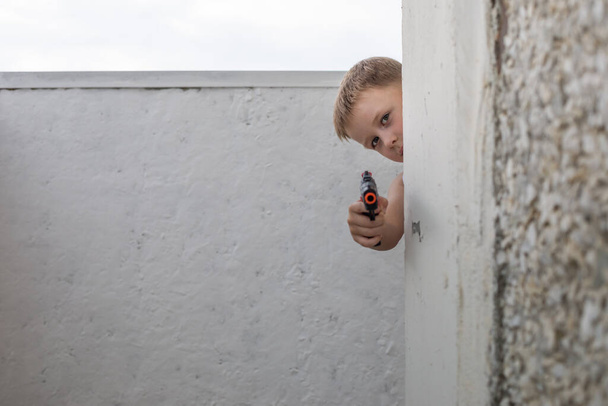 Маленький мальчик стреляет из игрушечного пистолета, прячась за углом здания
. - Фото, изображение