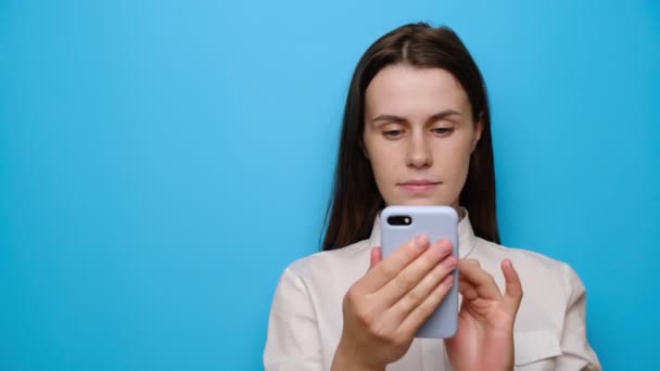 Portrait of beautiful calm brunette girl stands with mobile phone, wears white shirt, points aside with index finger on blank space, isolated on blue studio background. People, religion, technology - Filmati, video