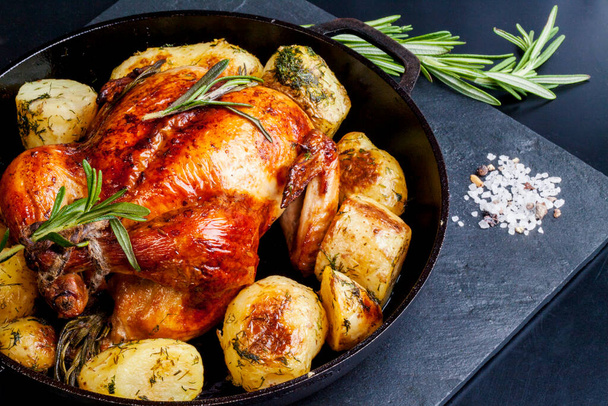 золотистый хрустящий курица на гриле в духовке с картофельными клинками и свежий розмарин в чугунной сковороде на черном фоне. вид сверху
 - Фото, изображение