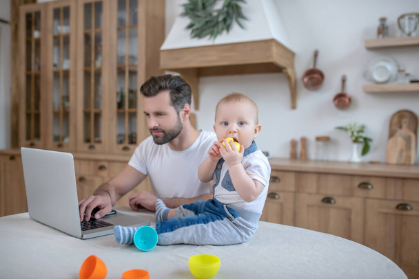 Dark-haired father working while his baby boy sitting on the table near him - Photo, image