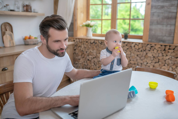 Dark-haired father working on a laptop, his little son sitting next to him - Φωτογραφία, εικόνα