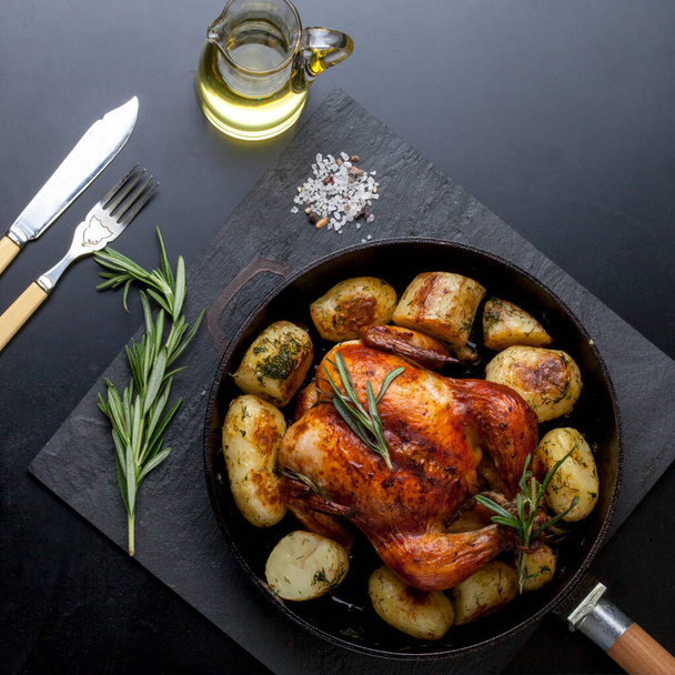 golden crispy grilled chicken in the oven with potato wedges and fresh rosemary in a cast-iron frying pan on a black background. view from above - Photo, Image