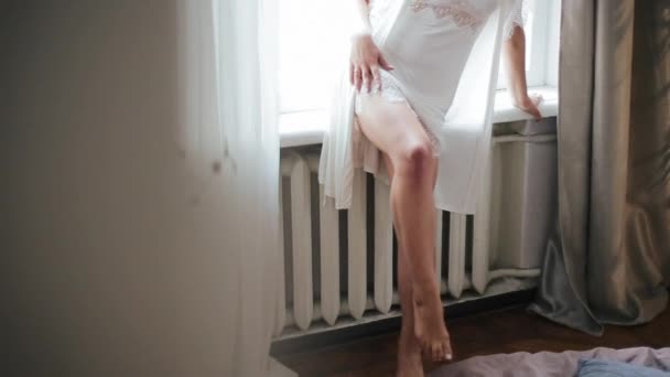 Krásné sexy nohy mladé evropské nevěsty v bílém peignoiru, opírající se ráno o parapet u okna pokoje. Pěkně usměvavá žena s hladkými elegantními nohami přehozenými přes sebe - Záběry, video