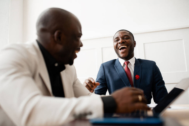 Twee zwarte zakenmannen lachen en discussiëren over wereldmarktnieuws terwijl ze aan een tafel zitten in een kantoor in pakken.. - Foto, afbeelding