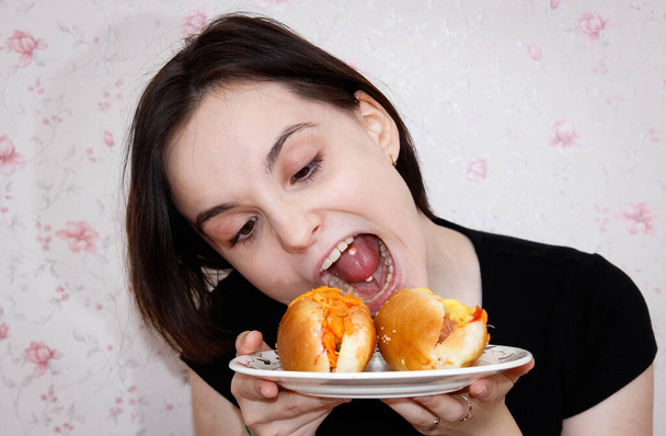 A girl with a piercing in her tongue and a grimace on her face tries to bite off a hot dog on a plate. Close-up, selective focus. - Photo, Image