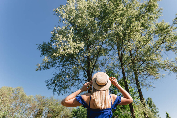 Portrait of a young woman wearing straw hat and blue dress standing back in rural landscape. Summer time, active life concept. - Photo, Image