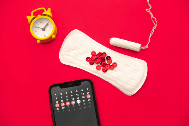 Cycle menstruel. Calendrier du mois avec des marques dans l'application mobile sur l'écran du smartphone. Alarme, tampon en coton, serviette hygiénique, étincelles rouges sur le fond rouge - Photo, image