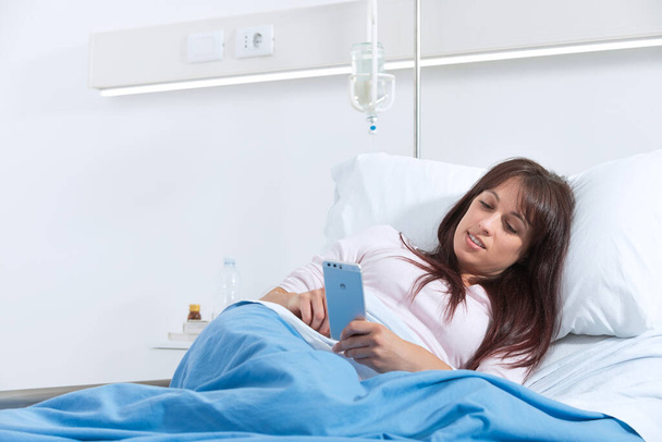 Sorridente paziente femminile sdraiata a letto in ospedale e chatta con il suo smartphone - Foto, immagini