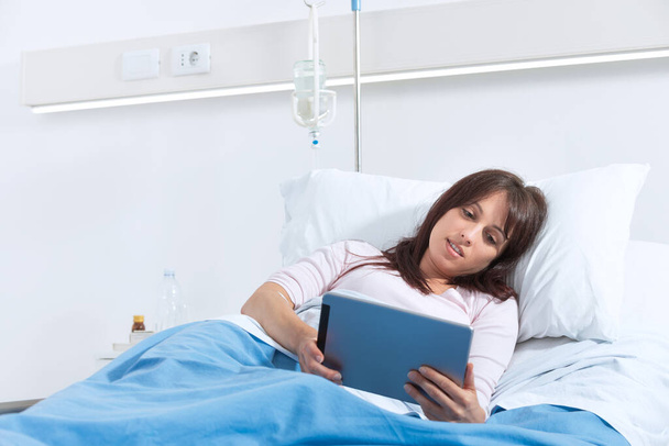 若いです笑顔患者は病院のベッドに横たわって、彼女はデジタルテーブルでビデオを見ています - 写真・画像