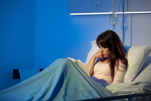 Young woman lying in a hospital bed at night and reading a book - Photo, Image