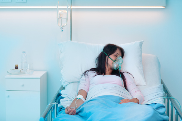 夜に病院のベッドに寝そべって酸素マスクをしている若い女性 - 写真・画像
