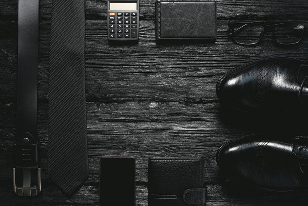 Business flat lay background with a copy space. Male shoes, belt, mobile phone, wallet, glasses and a necktie on black wooden floor. - Φωτογραφία, εικόνα