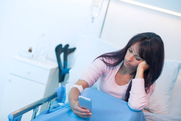 Gelangweilte Patientin liegt im Krankenhaus im Bett und chattet mit ihrem Smartphone - Foto, Bild
