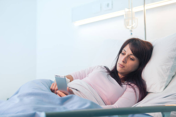 寝たきりの女性患者が病院のベッドに横になり、彼女のスマートフォンとチャット - 写真・画像