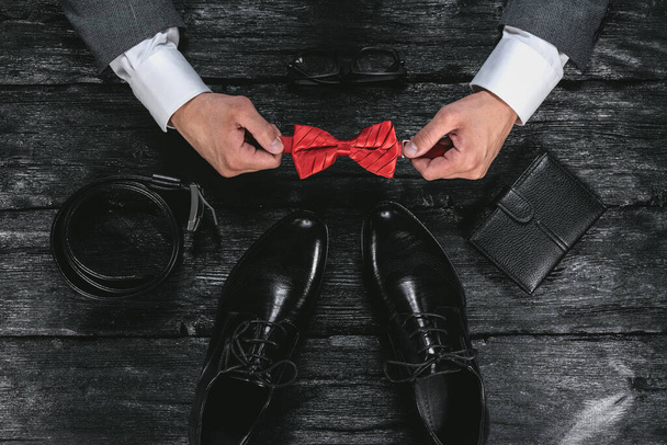 Business flat lay background. Male shoes, wallet, belt and a businessman hands with a red bow on black wooden table background. - Photo, image