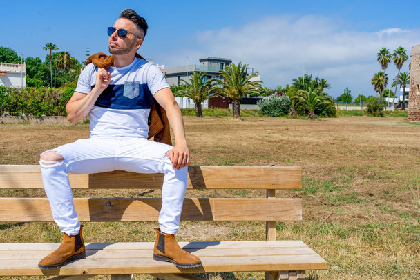 Nuori mies pukeutunut valkoisiin vaatteisiin ja moderni muotoilu poseeraa istuu puinen penkki. Muoti käsite - Valokuva, kuva