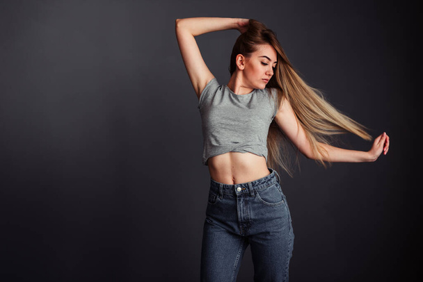 A thin girl in a gray top and jeans with blond long hair, hand behind head and looks away straightening her hair against a dark wall - Photo, image