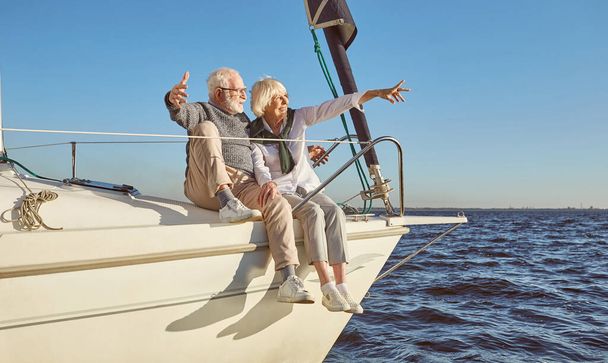 Een gelukkig senior echtpaar zit aan de zijkant van een zeilboot op een kalme blauwe zee, wijzend naar landschap, genietend van uitzicht - Foto, afbeelding