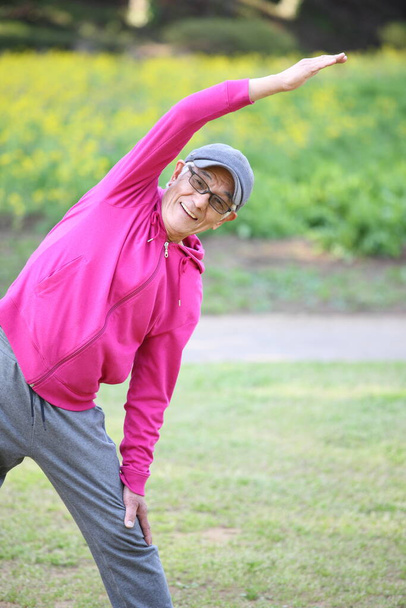 senior Japanese man wearing pink parka doing standing side bend exercise - Фото, изображение