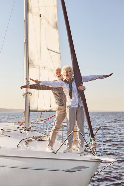 Against the wind. Happy senior couple standing on the side of sail boat or yacht deck floating in sea. Man and woman stretching hands out against the sky and smiling - Photo, Image