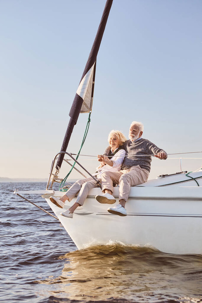 A happy senior couple talking, sitting on the side of a sail boat on a calm blue sea. Man hugging his woman while enjoying view - Photo, Image
