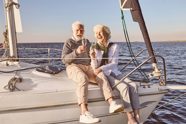 Joyeux couple de personnes âgées assis sur le côté du voilier ou du pont de yacht flottant en mer. Homme et femme buvant du vin ou du champagne et riant - Photo, image