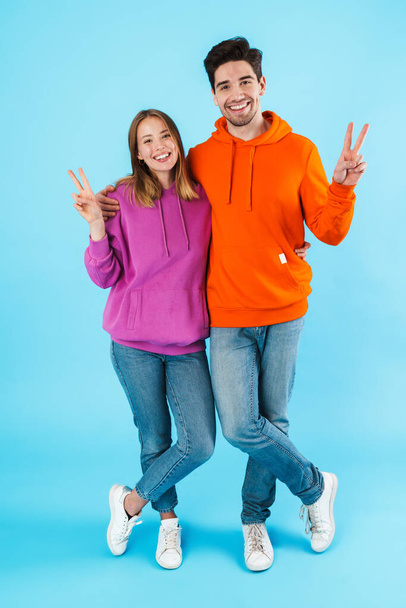 Full length portrait of a cheerful young couple wearing hoodies standing isolated over blue background, hugging, peace gesture - Фото, зображення