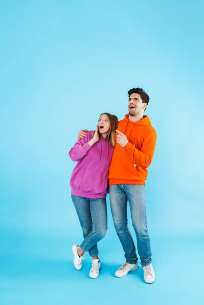 Full length portrait of a cheerful young couple wearing hoodies standing isolated over blue background, looking away at copy space - Fotó, kép
