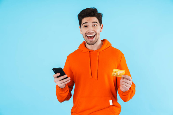 Portrait of a cheerful young man wearing hoodie standing isolated over blue background, holding mobile phone, using credit card, paying - Fotografie, Obrázek