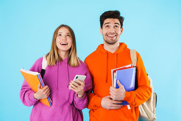 Porträt eines fröhlichen jungen Studentenpaares mit Rucksäcken und Schulbüchern, die isoliert vor blauem Hintergrund stehen und nach oben schauen - Foto, Bild