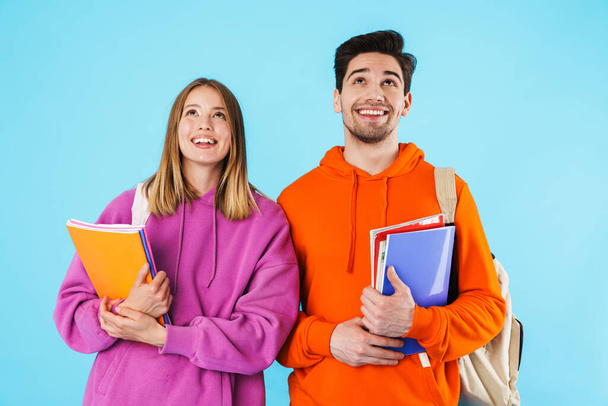Portrait of a cheerful young couple of students wearing backpacks, carrying textbooks standing isolated over blue background, looking up - Foto, Bild