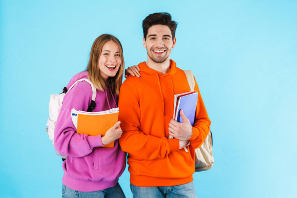 Portrait of a cheerful young couple of students wearing backpacks, carrying textbooks standing isolated over blue background - Fotografie, Obrázek