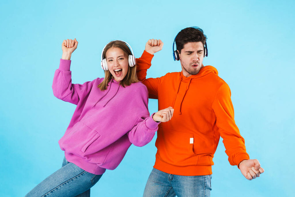 Portrait of a cheerful young couple wearing hoodies standing isolated over blue background, listening to music with wireless headphones, dancing - Fotó, kép