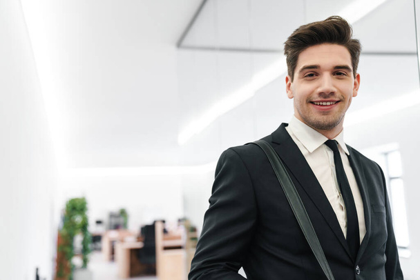 Образ счастливого молодого бизнесмена в черном костюме, улыбающегося стоя с сумкой в офисе
 - Фото, изображение