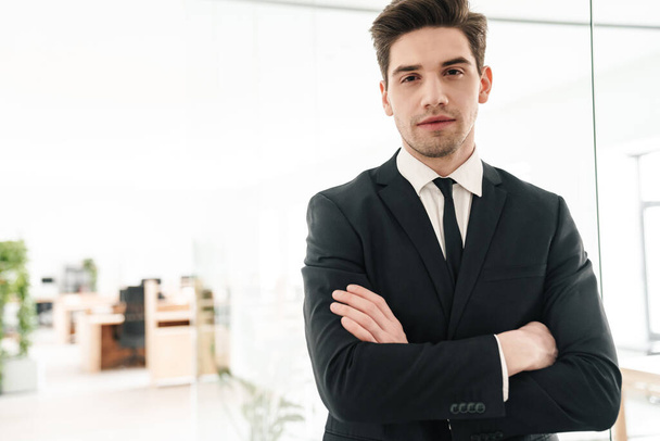 Image of serious young businessman wearing black suit standing with hands crossed in office - Foto, Imagen
