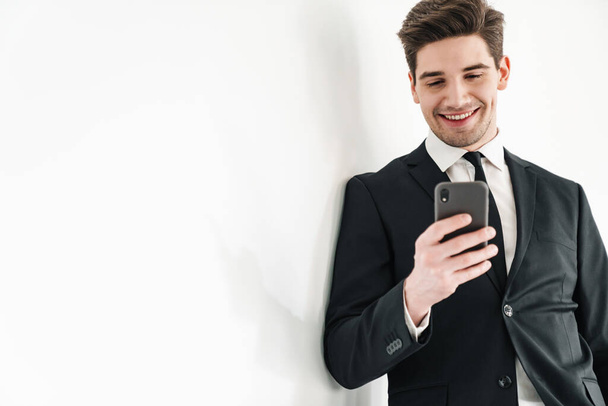 Image of smiling young businessman wearing black suit using mobile phone in office - Φωτογραφία, εικόνα