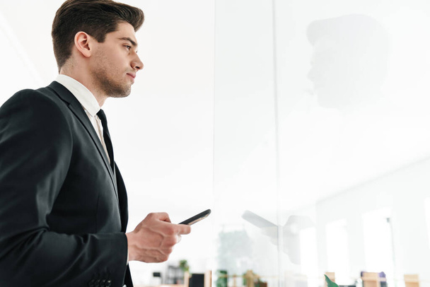 Image of serious young businessman wearing black suit using mobile phone in office - Photo, Image