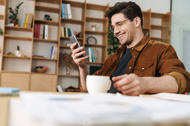 Image of handsome joyful man in eyeglasses smiling and using cellphone while drinking coffee at office - Foto, imagen