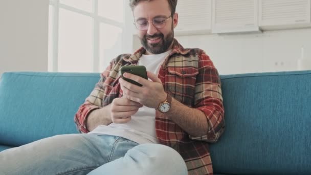 Handsome young positive happy bearded man indoors at home sitting on a sofa while using mobile phone - Metraje, vídeo