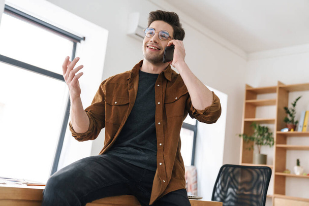 Image of handsome joyful young man smiling and talking cellphone while working in office - Foto, afbeelding
