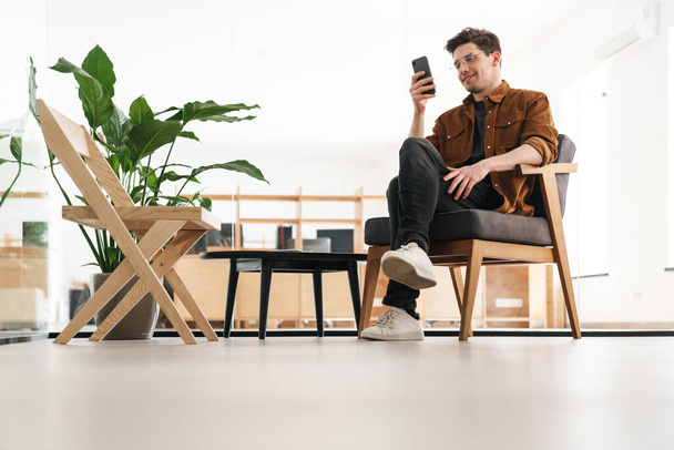 Image of handsome joyful young man smiling and using cellphone while working in office - Fotoğraf, Görsel