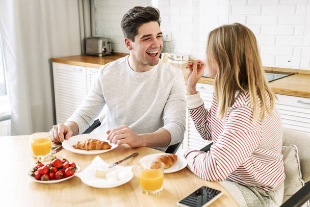 Portrait of young happy couple eating together at table while having breakfast in cozy kitchen at home - Foto, immagini