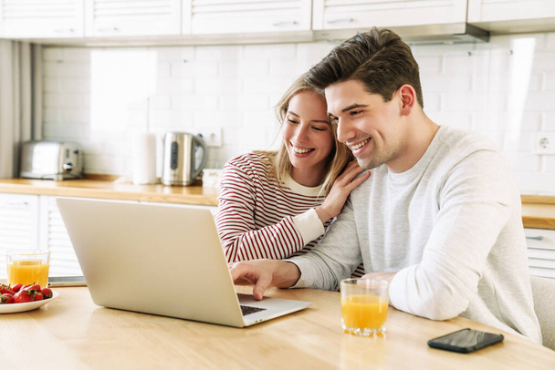 Portrait of young cheerful couple using laptop and smiling while having breakfast in cozy kitchen at home - Foto, immagini