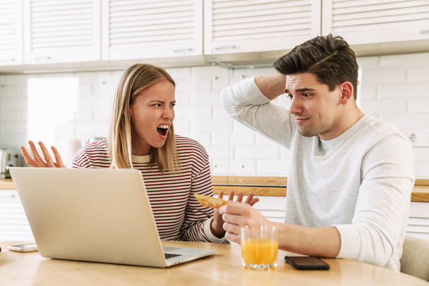 Portrait of displeased couple using laptop and holding credit card while sitting in kitchen at home - Foto, afbeelding