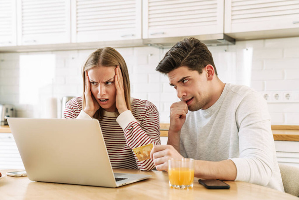 Portrait of displeased couple using laptop and holding credit card while sitting in kitchen at home - Foto, Bild