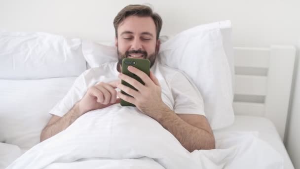 Handsome young pleased bearded man indoors at home lies in bed while using smartphone - Felvétel, videó