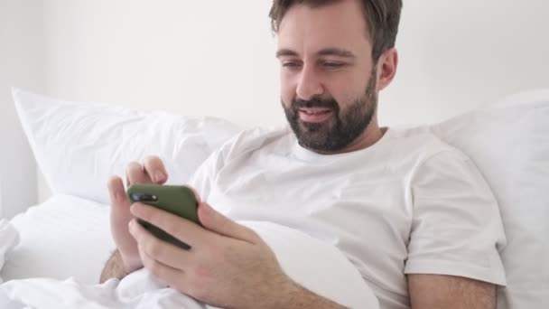 Handsome young concentrated bearded man indoors at home lies in bed while using smartphone - Séquence, vidéo